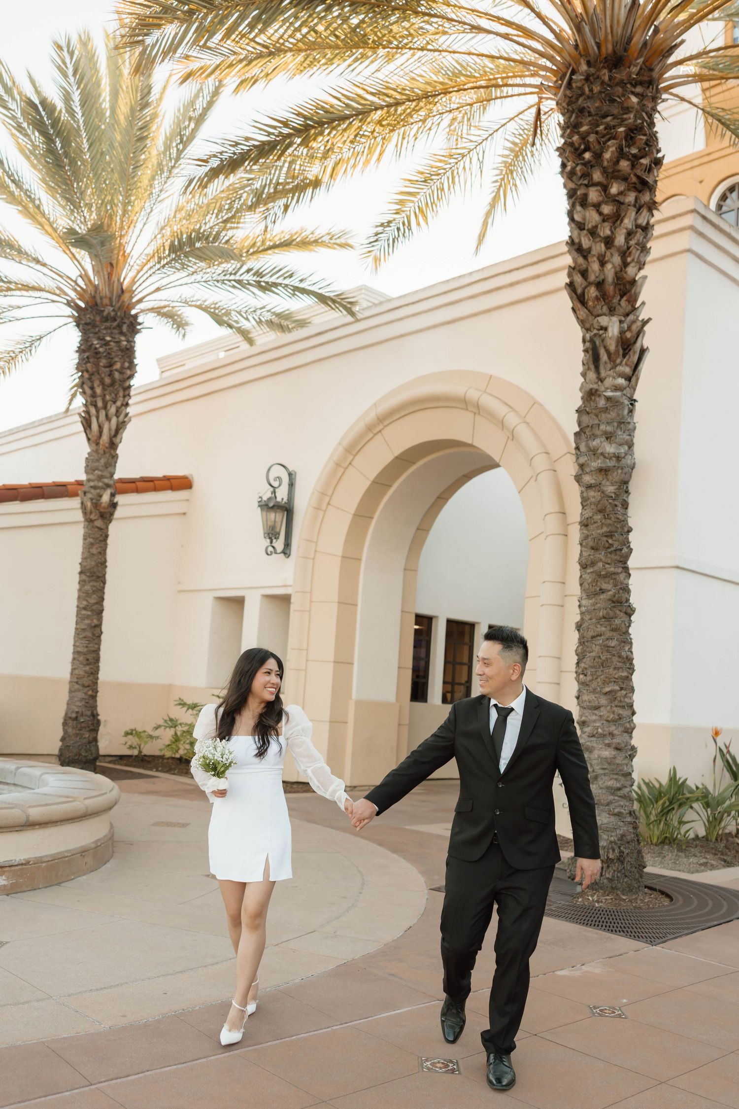 A couple walks along a Mediterranean-style walkway with palm trees and arched architecture in an elegant setting.