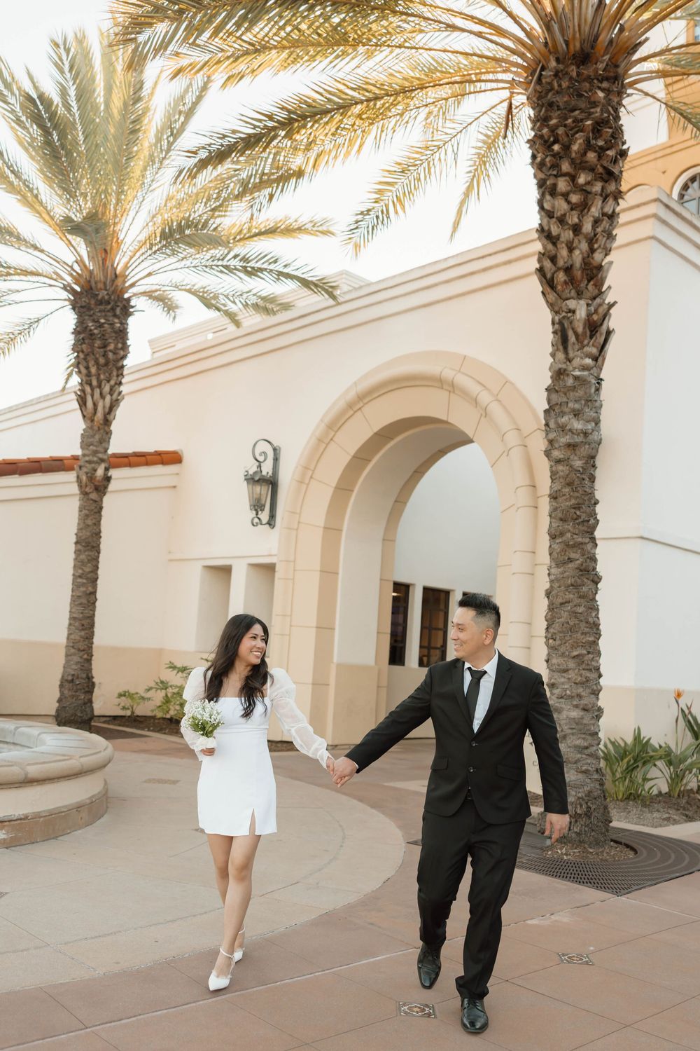 A couple walks along a Mediterranean-style walkway with palm trees and arched architecture in an elegant setting.