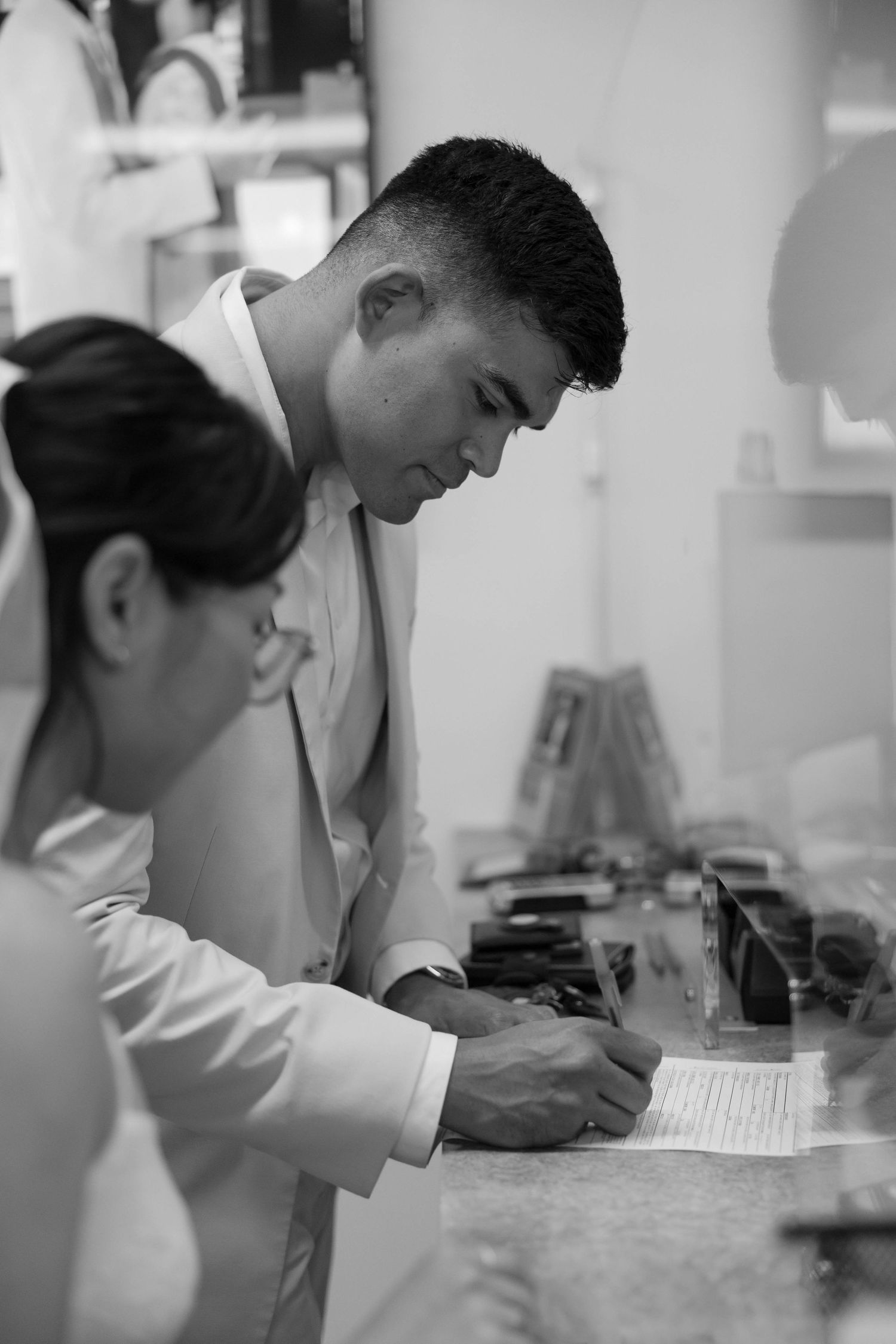 Black and white photo of a man in a white suit signing a document at a counter beside a woman.