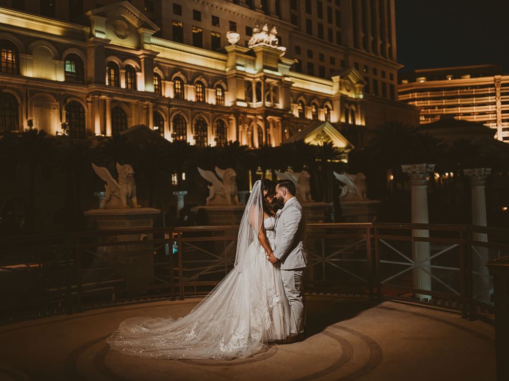 A romantic wedding couple holds each other close on a dark, illuminated bridge with elegant city lights in the background.