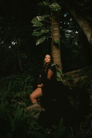 Woman in black dress and boots poses among lush tropical foliage in a dark, moody forest setting.