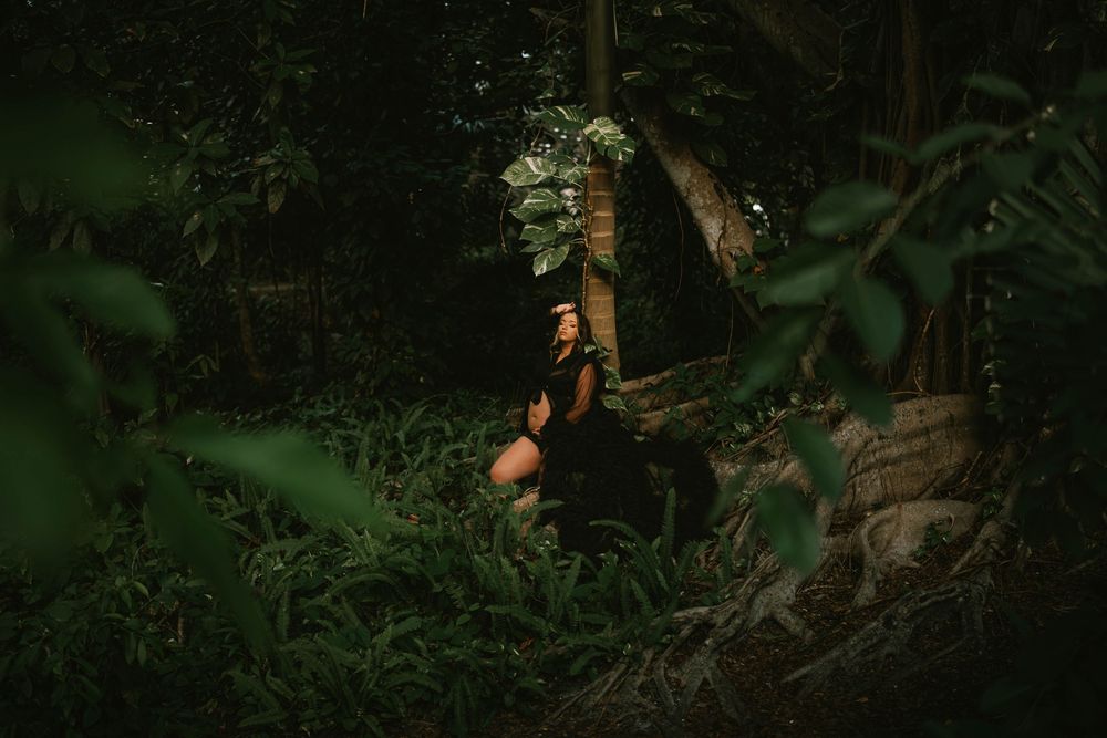 A woman in black dress crouches in a lush tropical jungle surrounded by dense green foliage and trees.