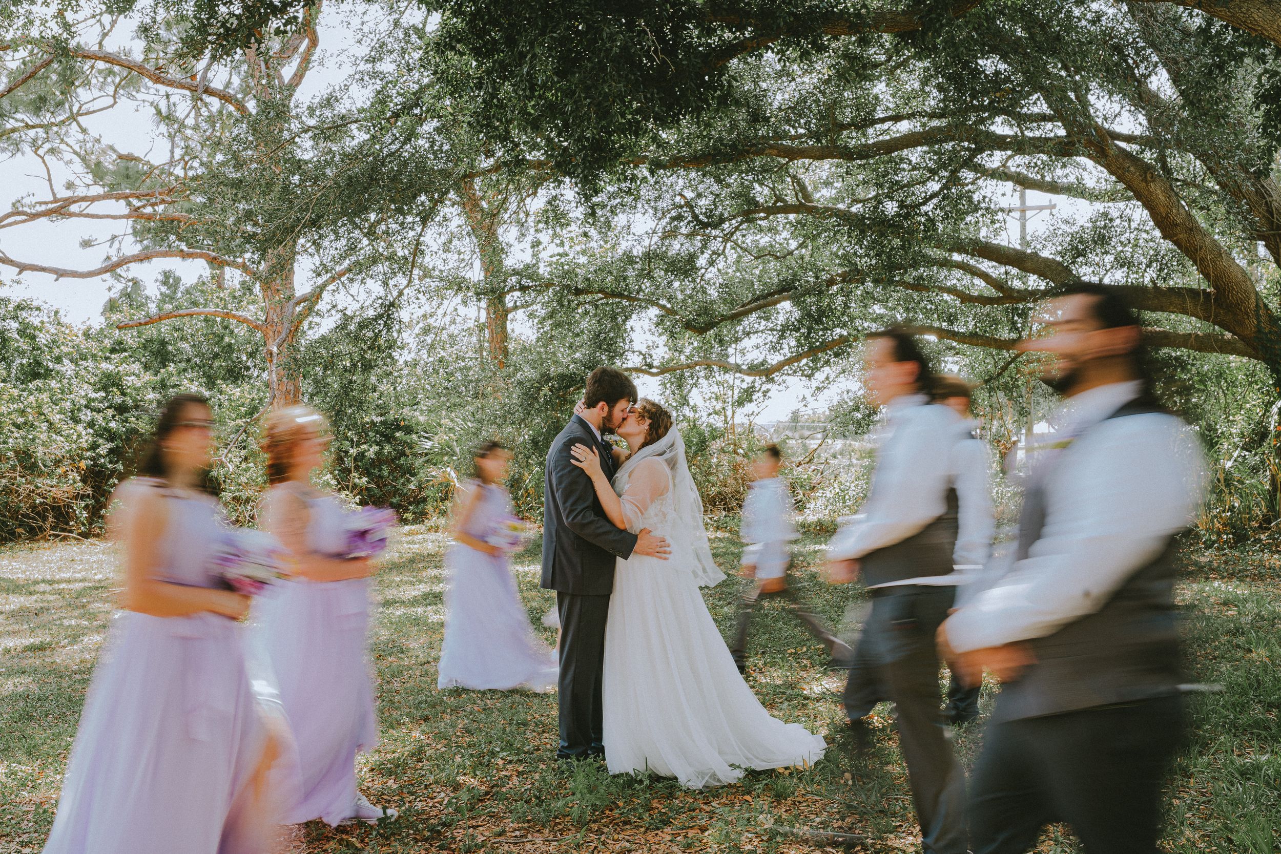 Wedding party in purple dresses dancing in blurred motion under trees during outdoor celebration.