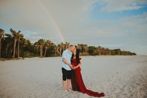 Couple kissing on white sand beach with rainbow and palm trees, pregnant woman in flowing red maternity gown.