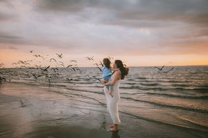 Woman holding child on beach at sunset with seagulls flying over ocean waves in golden evening light.