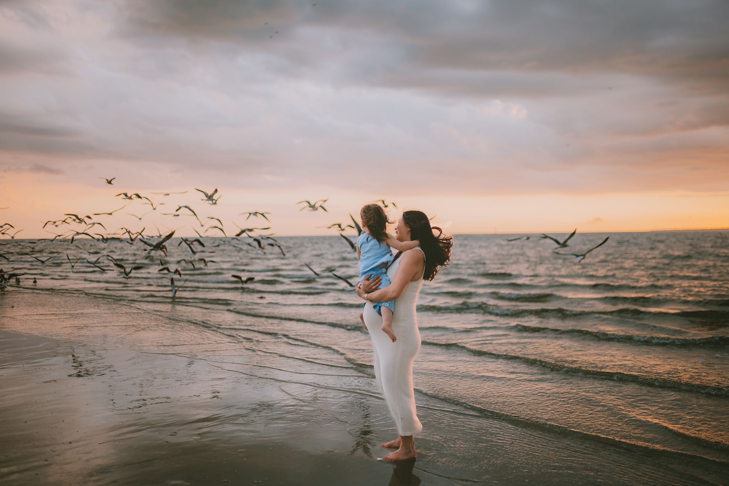 Woman holding child on beach at sunset with seagulls flying over ocean waves in golden evening light.