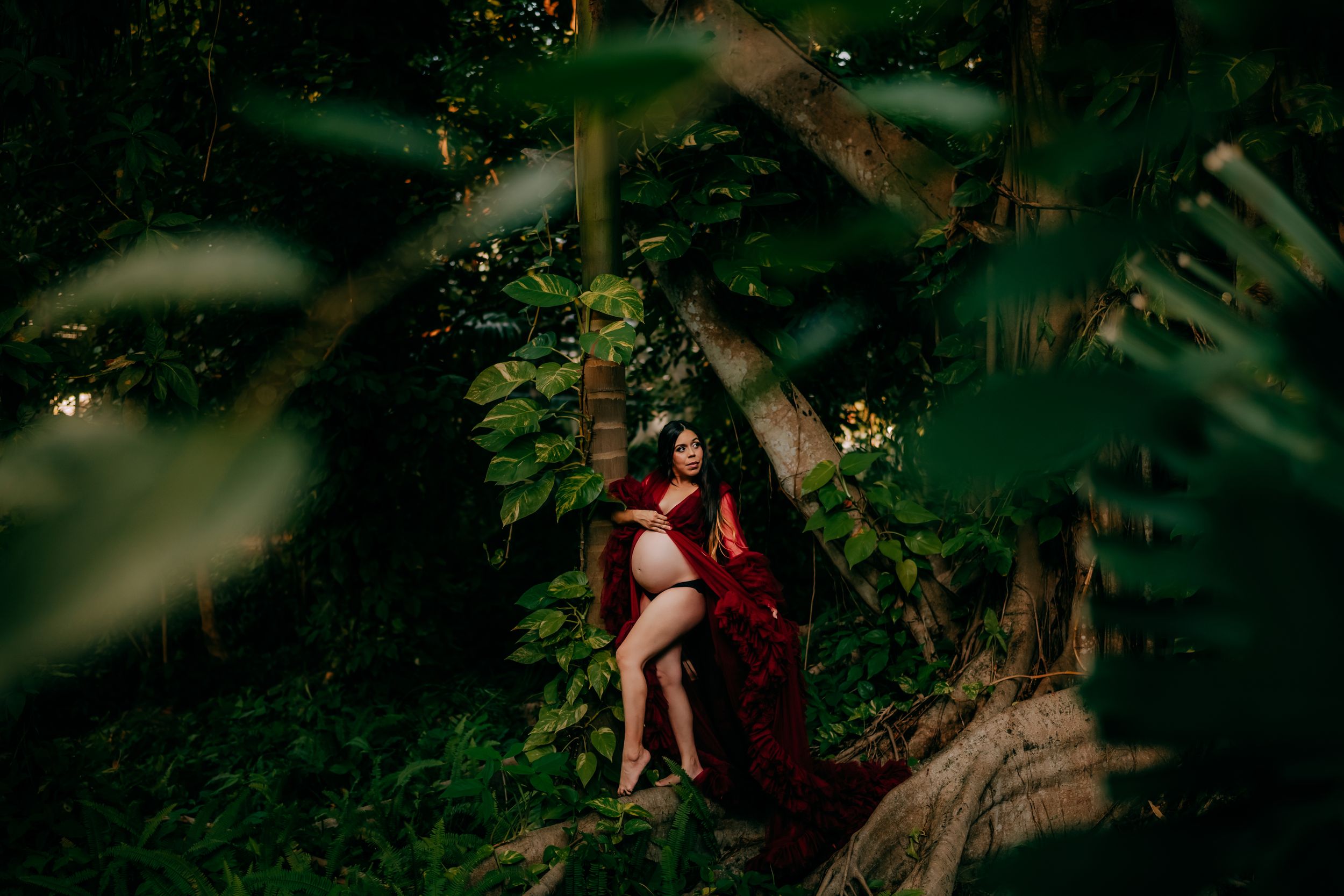 A pregnant woman in a flowing red gown poses gracefully among lush green trees in a serene outdoor setting.