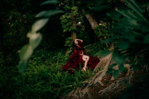 A woman in a flowing red gown sits gracefully among lush green forest foliage in a dramatic editorial photo.