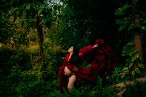 Pregnant woman in flowing red gown poses dramatically among lush green forest trees in a stunning maternity photo.