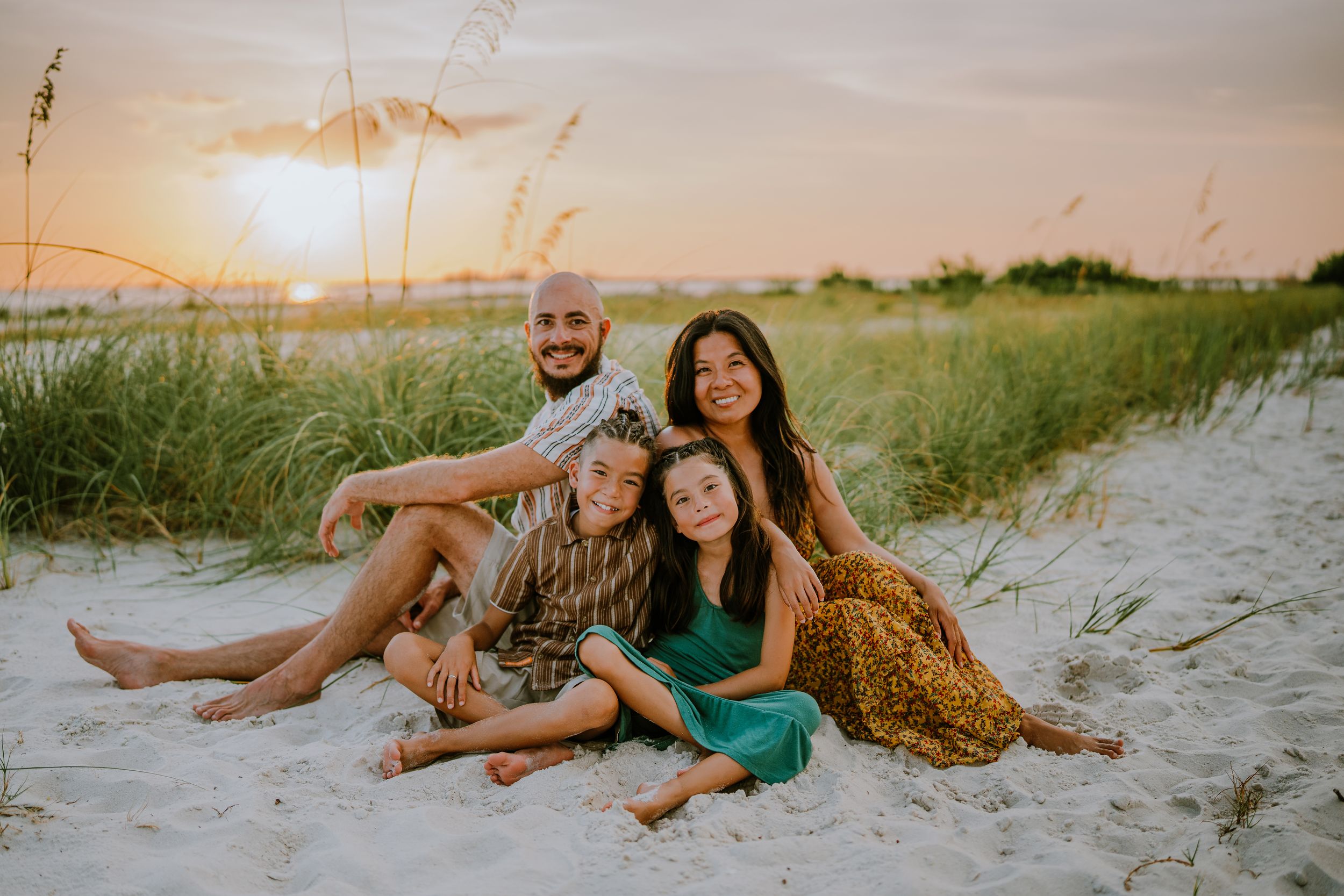 Family of four smiling together on sandy beach at sunset with sea oats in background.