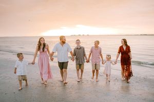 Family of seven holding hands walking on a beach at sunset, wearing coordinated outfits in pink, white, and burgundy.
