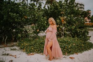 Pregnant woman in pink off-shoulder maxi dress poses on white sand beach surrounded by lush green tropical trees.