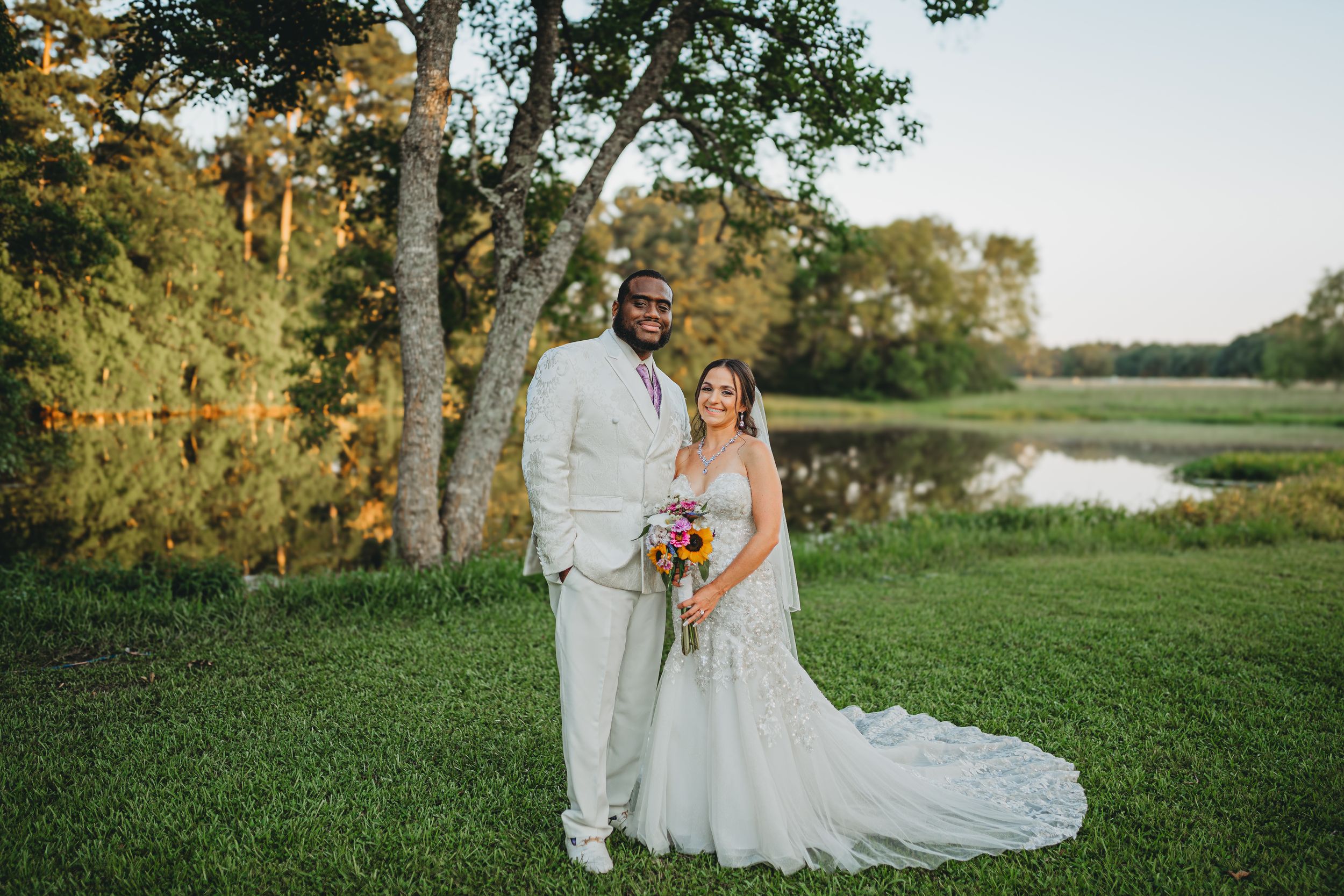 Couple stands together outdoors on grassy lawn near birch trees and pond during sunset wedding ceremony.