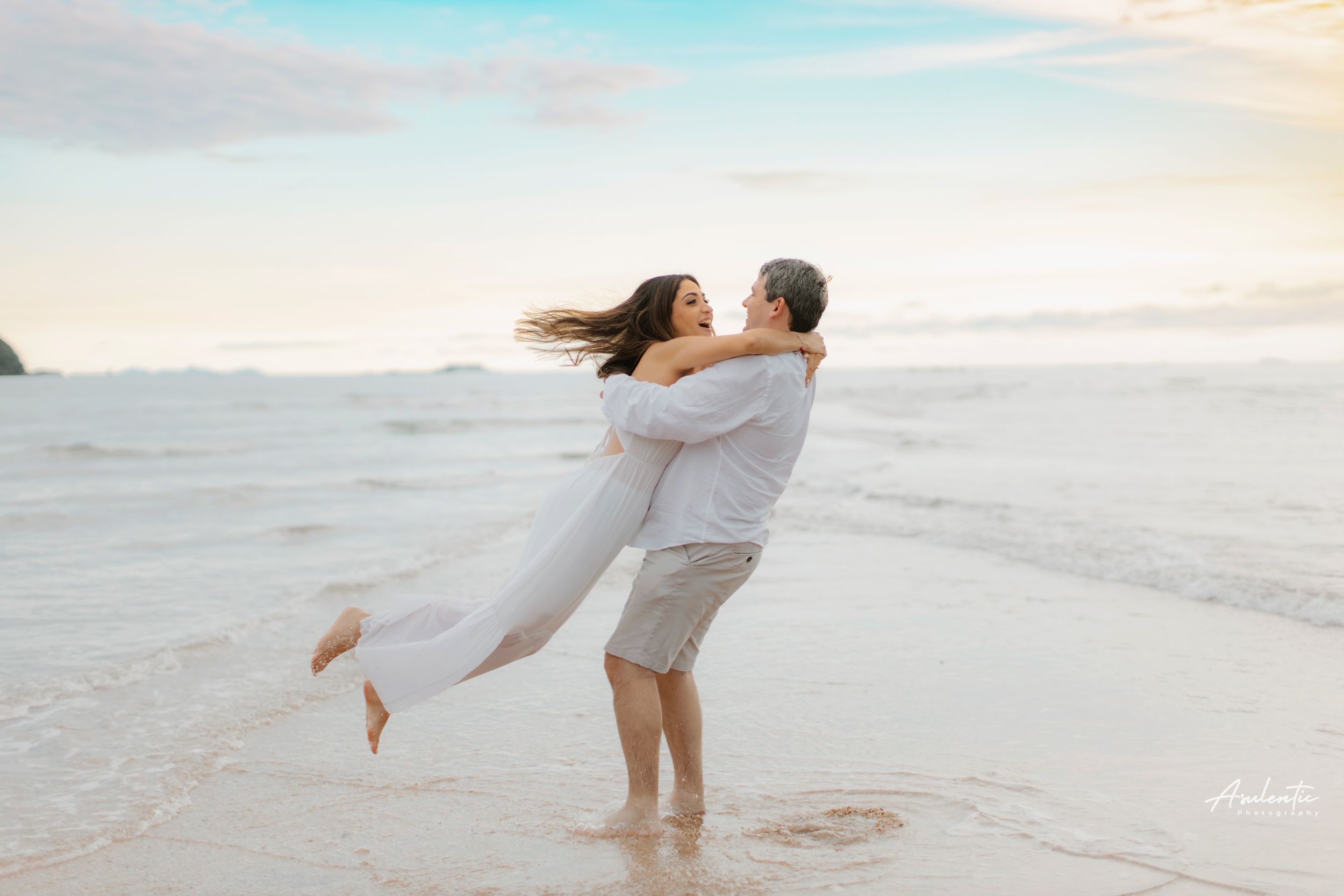 Romantic couple in light clothing embrace and spin on sandy beach at sunset.
