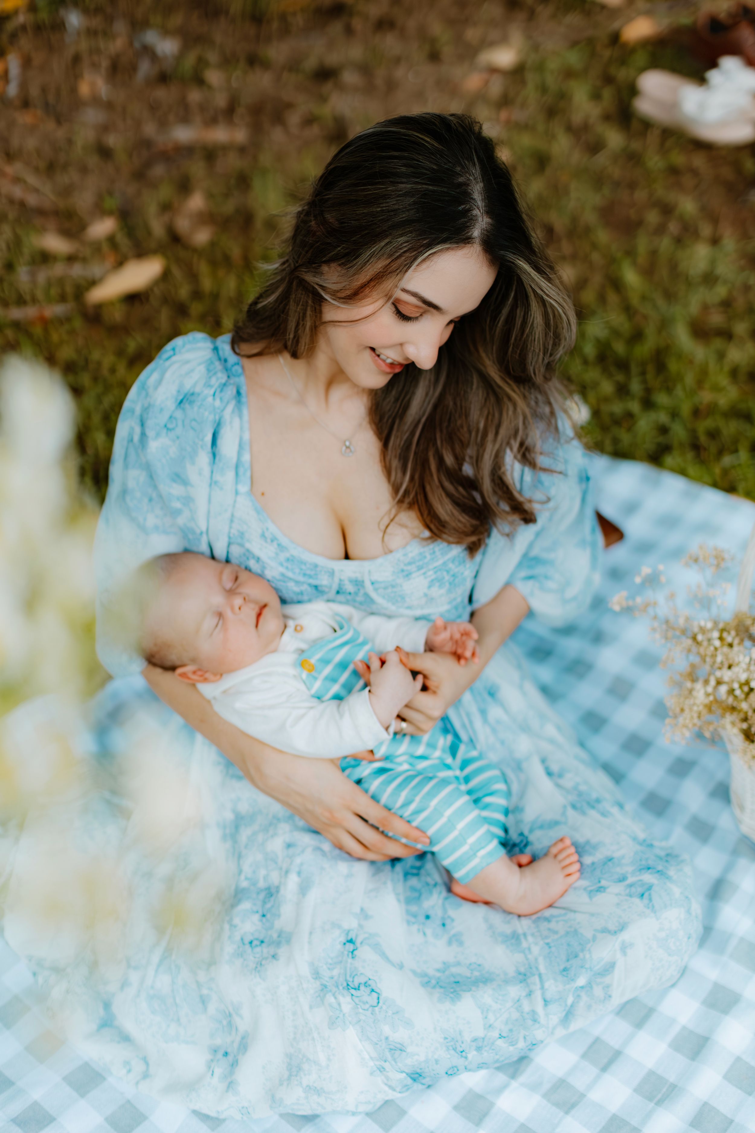 Mother in blue floral dress holding sleeping baby on picnic blanket outdoors.