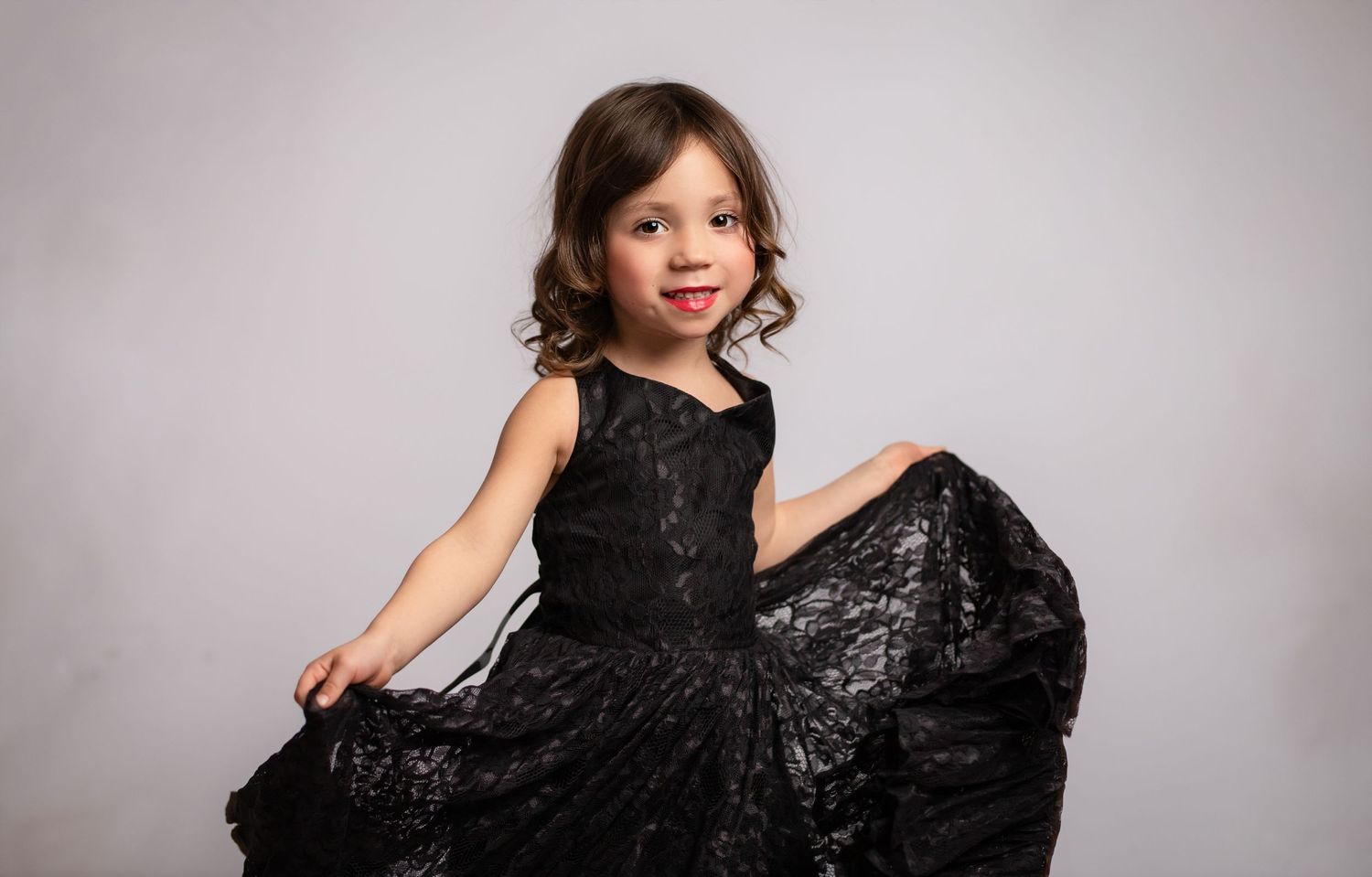 A child in an elegant black lace dress twirls playfully against a gray studio backdrop.