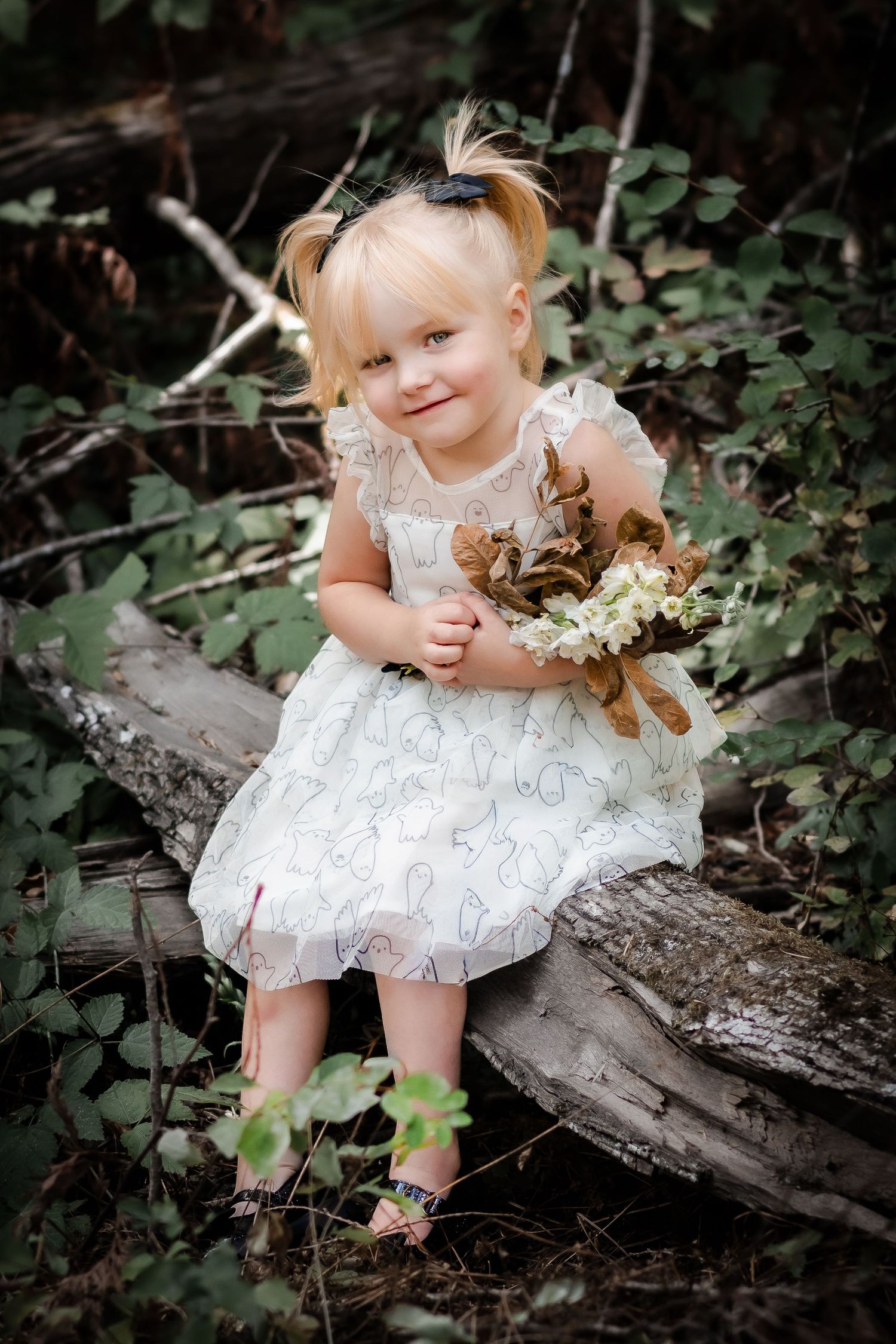 Blonde toddler girl in white lace dress sits on log holding flowers, smiling in a lush green forest setting.