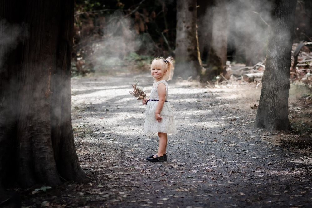 A young blonde girl in a white dress stands on a forest path surrounded by misty trees.