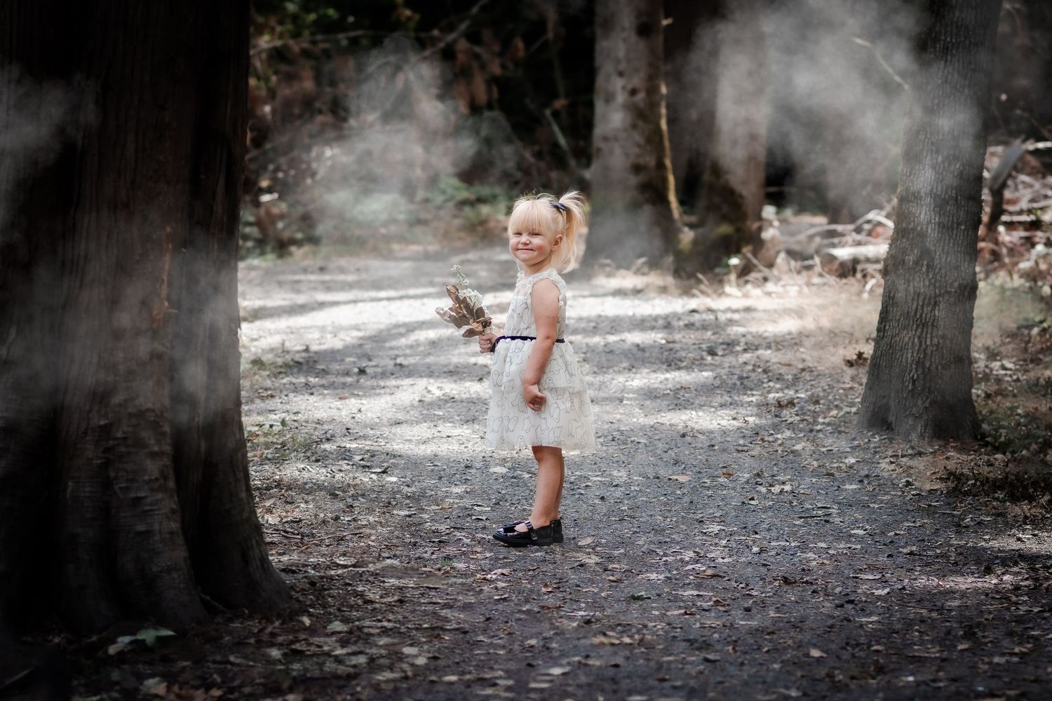 A young blonde girl in a white dress stands on a forest path surrounded by misty trees.