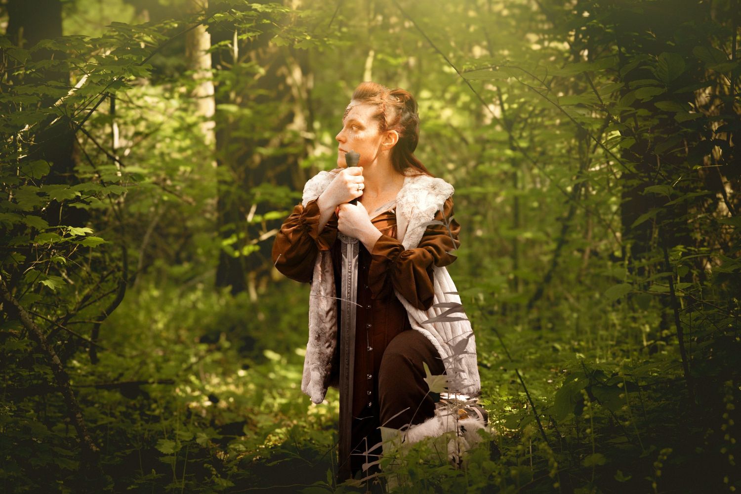 A woman in medieval costume stands in a sunlit forest, holding a sword with lush green trees surrounding her.