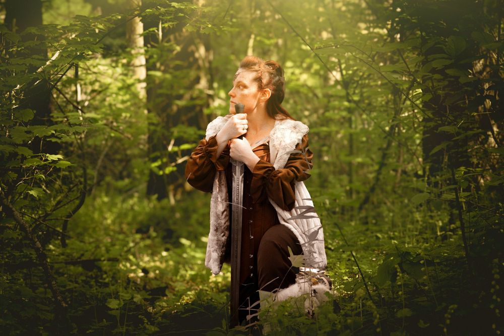 A woman in medieval costume stands in a sunlit forest, holding a sword with lush green trees surrounding her.