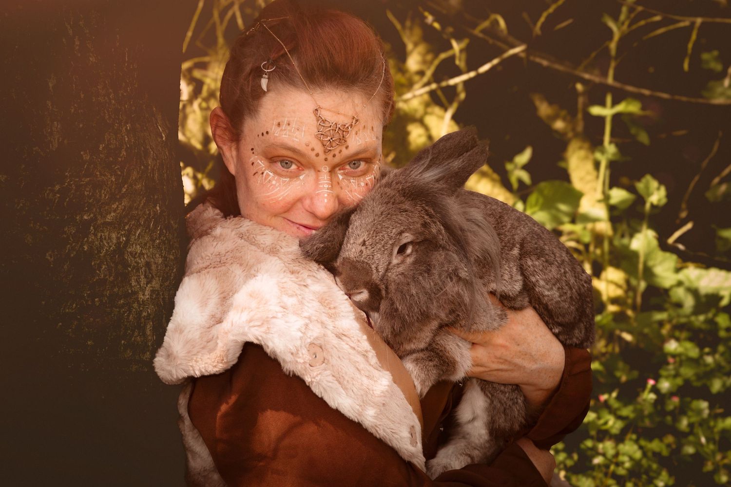 Woman with butterfly face paint tenderly holds a fluffy gray rabbit outdoors in golden light.