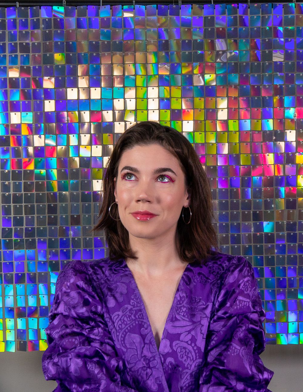 A female model poses against a sparkly mosaic backdrop wearing a purple velvet top.