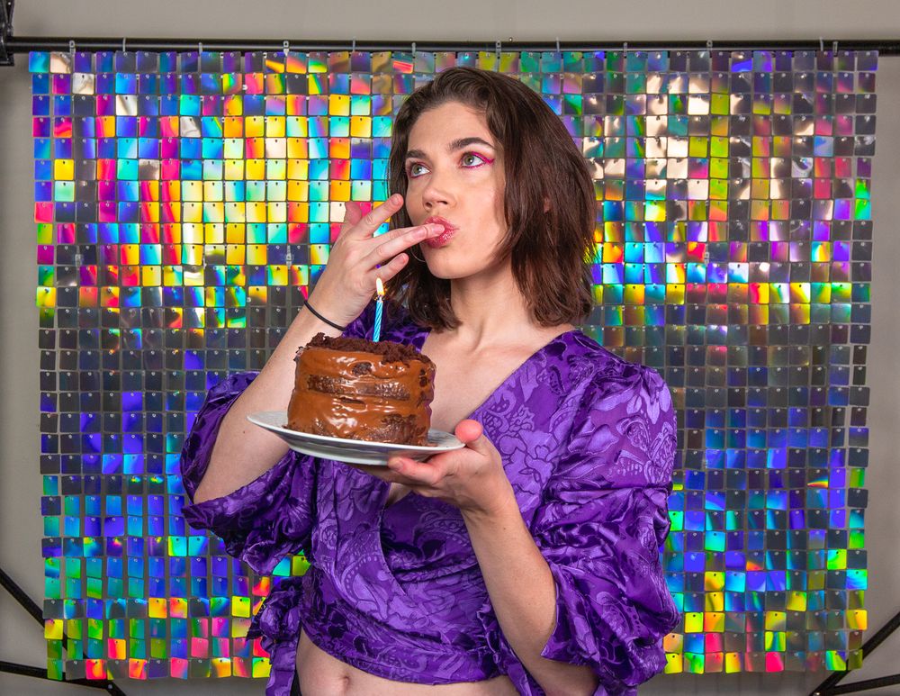 Woman in purple top enjoys chocolate cake against colorful mosaic background.