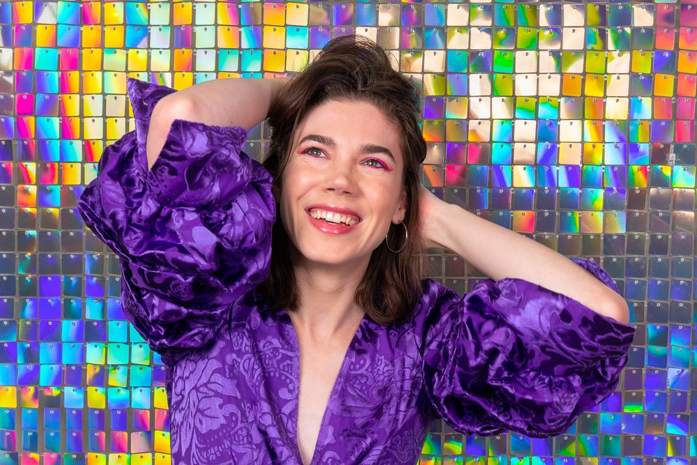 Woman in purple velvet top laughs joyfully against colorful mosaic background.