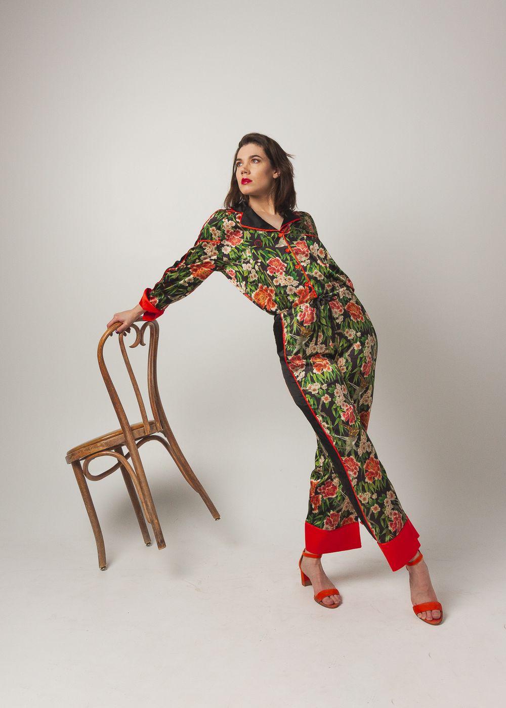 Model in floral print jumpsuit poses elegantly with vintage wooden chair.