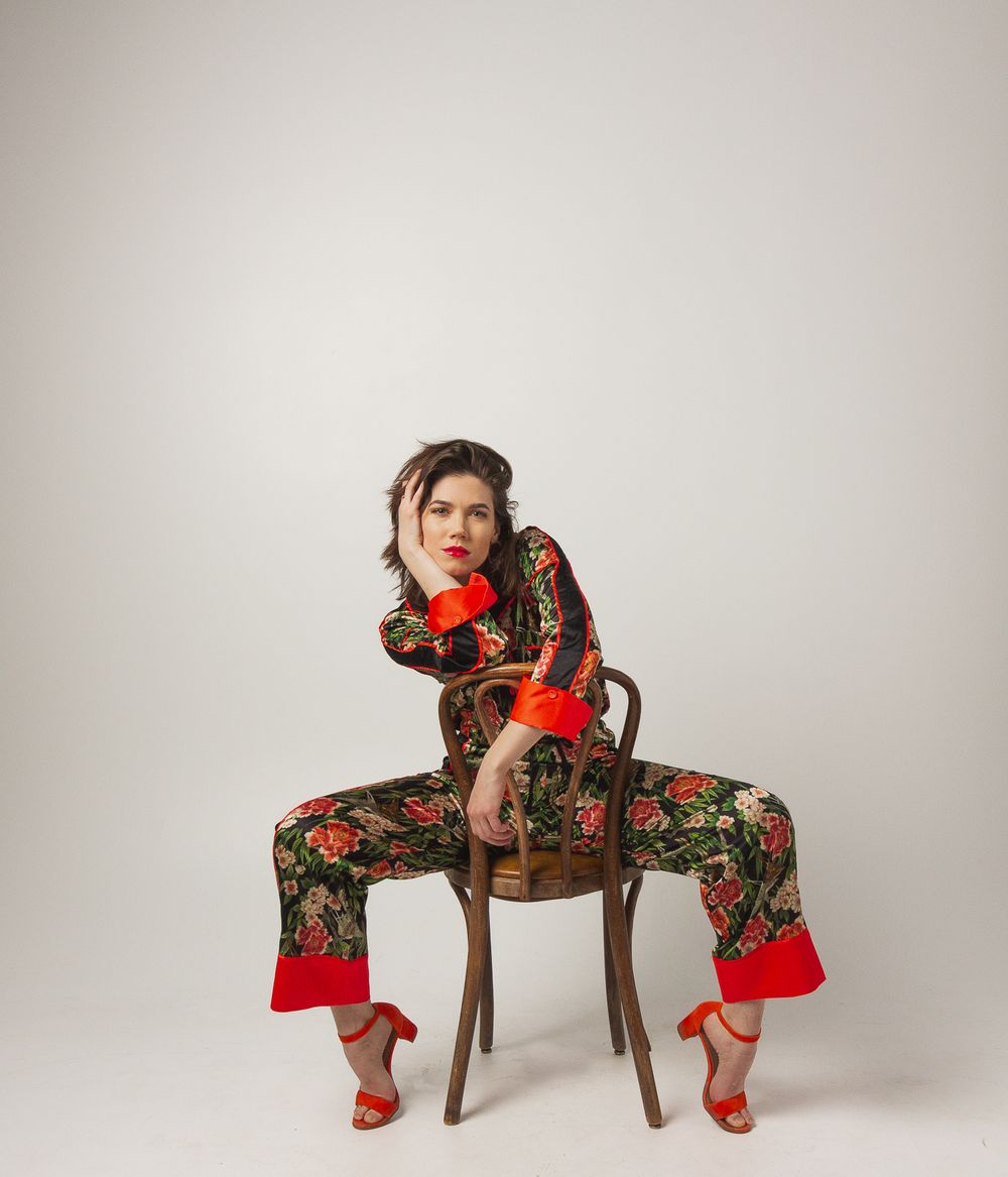 Model in floral jumpsuit with red accents poses on wooden chair against white background.