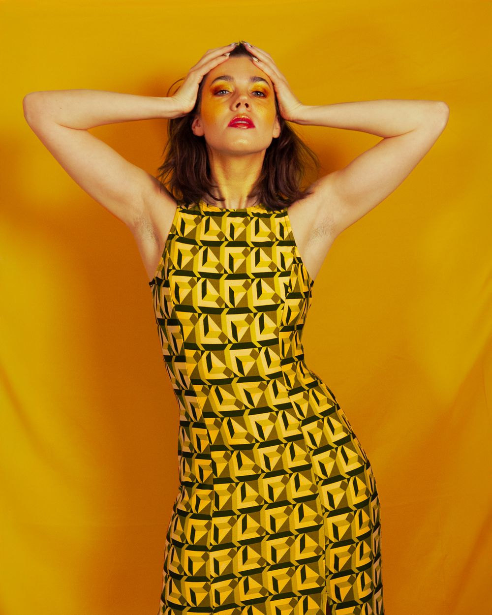 Model in yellow geometric print dress poses with hands on head against bright orange backdrop.