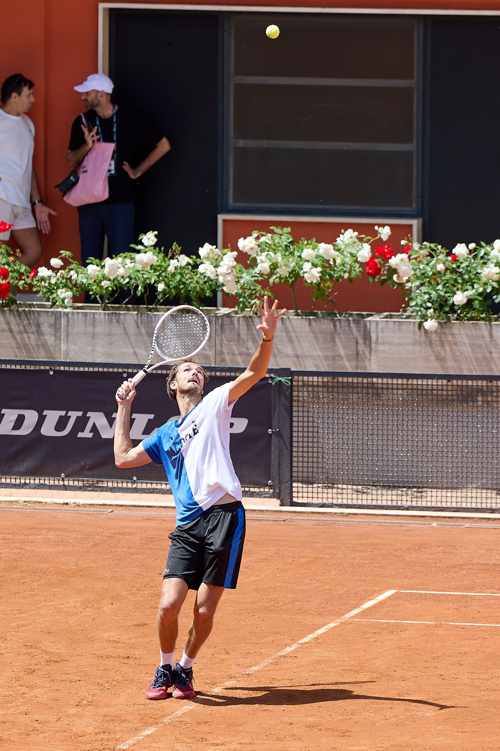 Tennis player serving on clay court with decorative flowers in background.