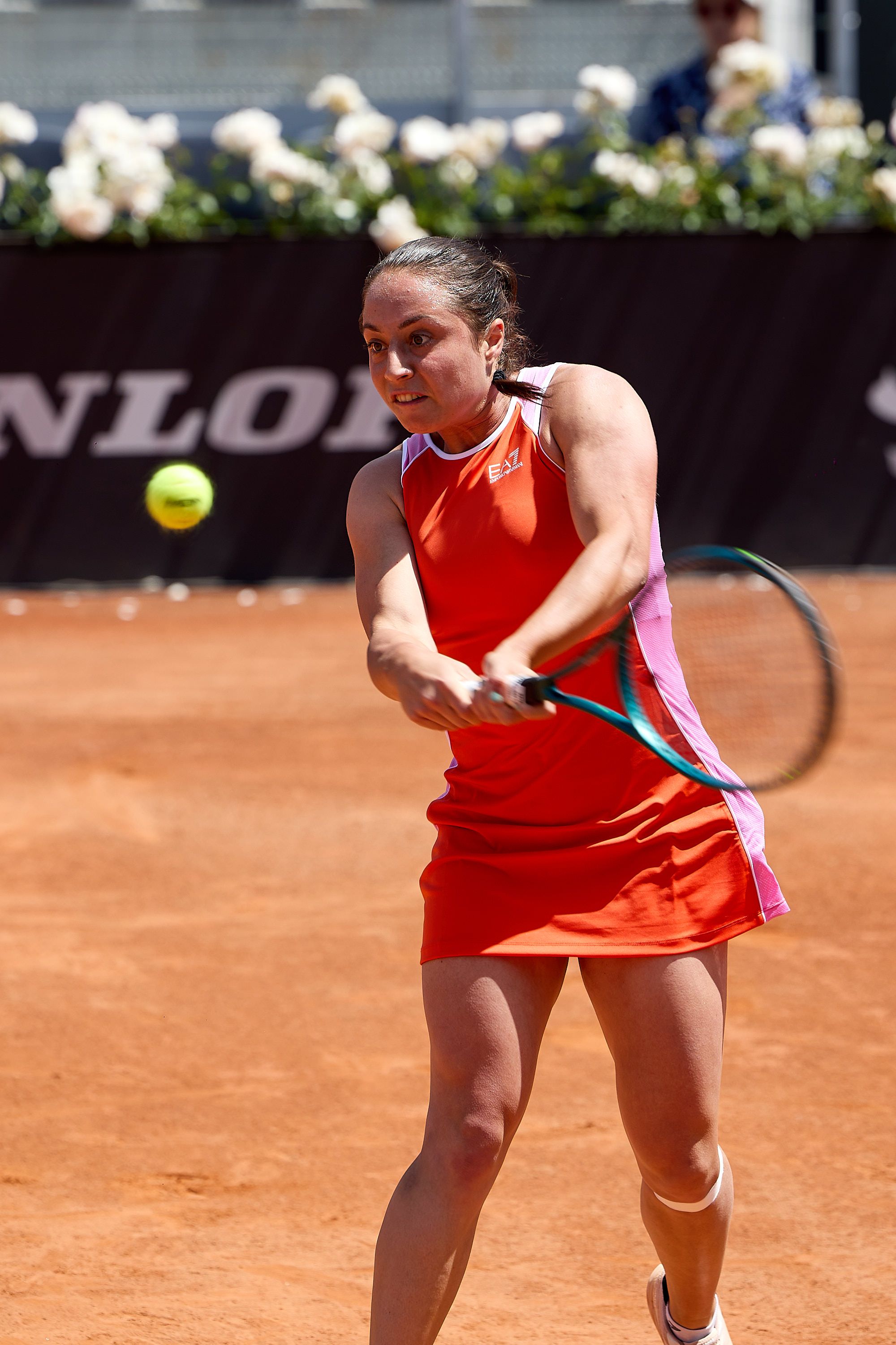 Female tennis player in red dress hits backhand shot on clay court during tournament match.