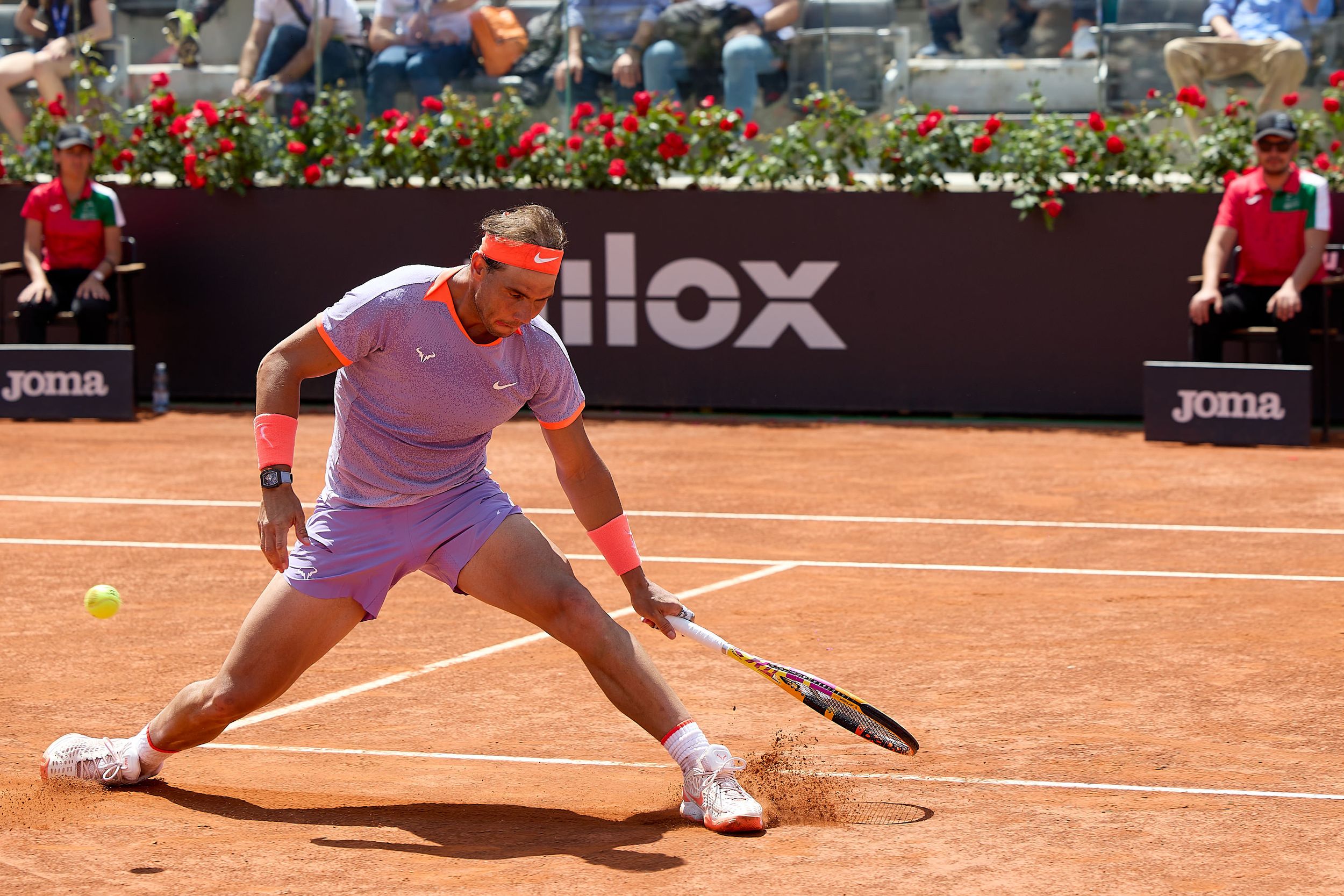 Tennis player stretches low to return a shot on clay court with red flowers in background at professional tournament.