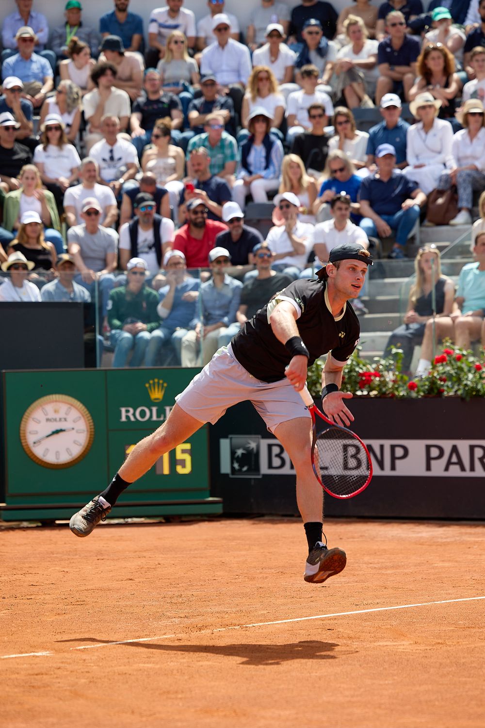 Tennis player serves ball on clay court with crowd and Rolex clock visible in background.