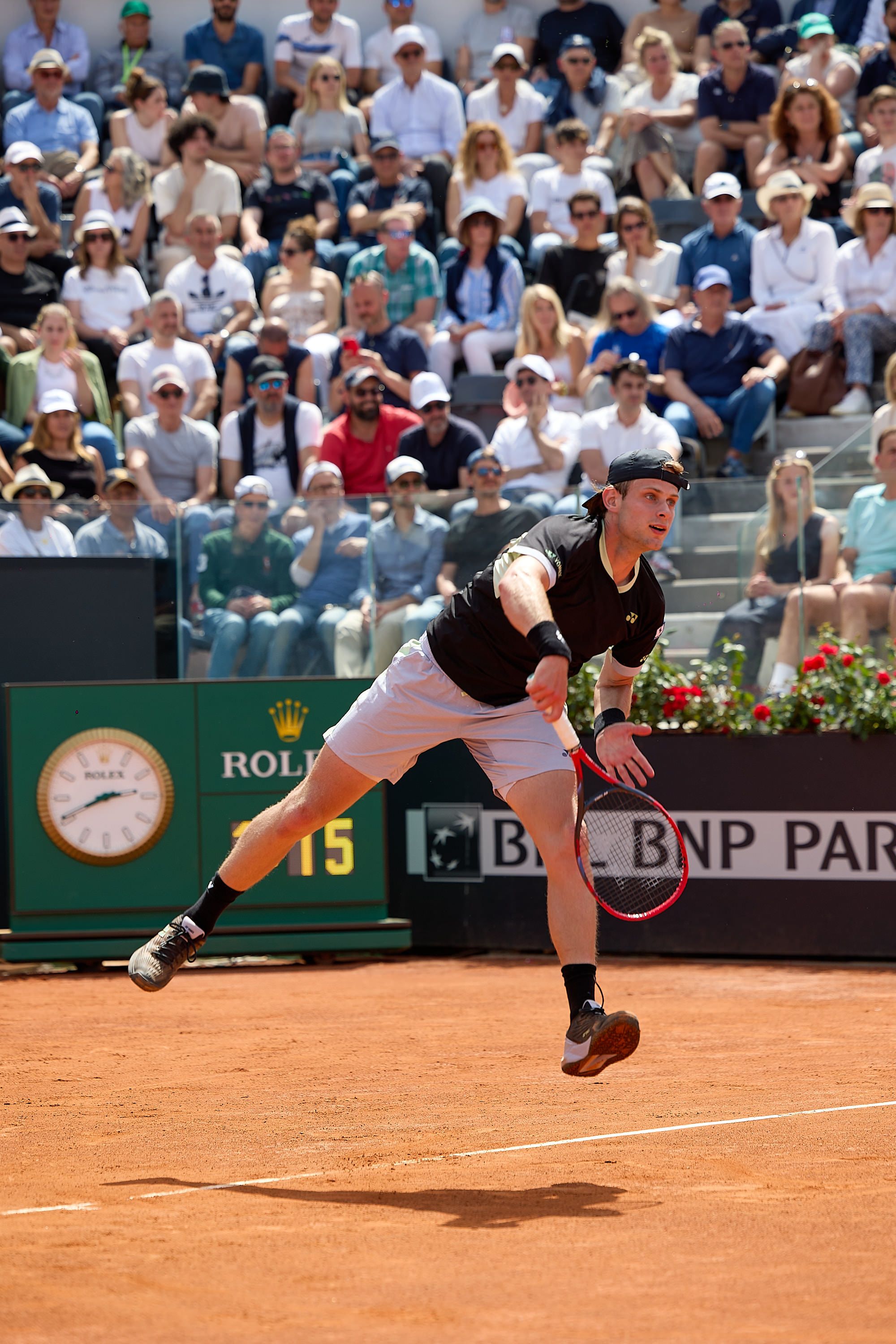 Tennis player serves ball on clay court with crowd and Rolex clock visible in background.
