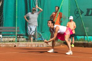 Tennis players practicing serves and returns on clay court with green fence background.