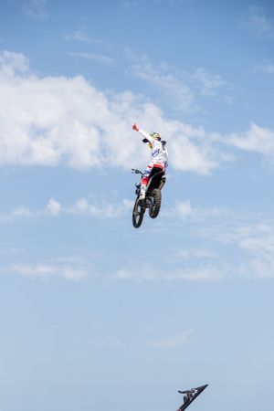 Motocross rider performs dramatic aerial jump against blue sky and white clouds.