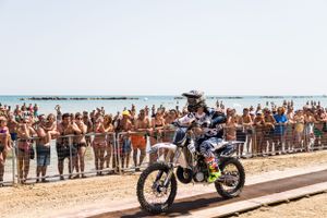 Dirt bike rider races past cheering crowd on beach during motorsport event.