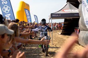 Motorcycle rider races through crowd of spectators along beach course.