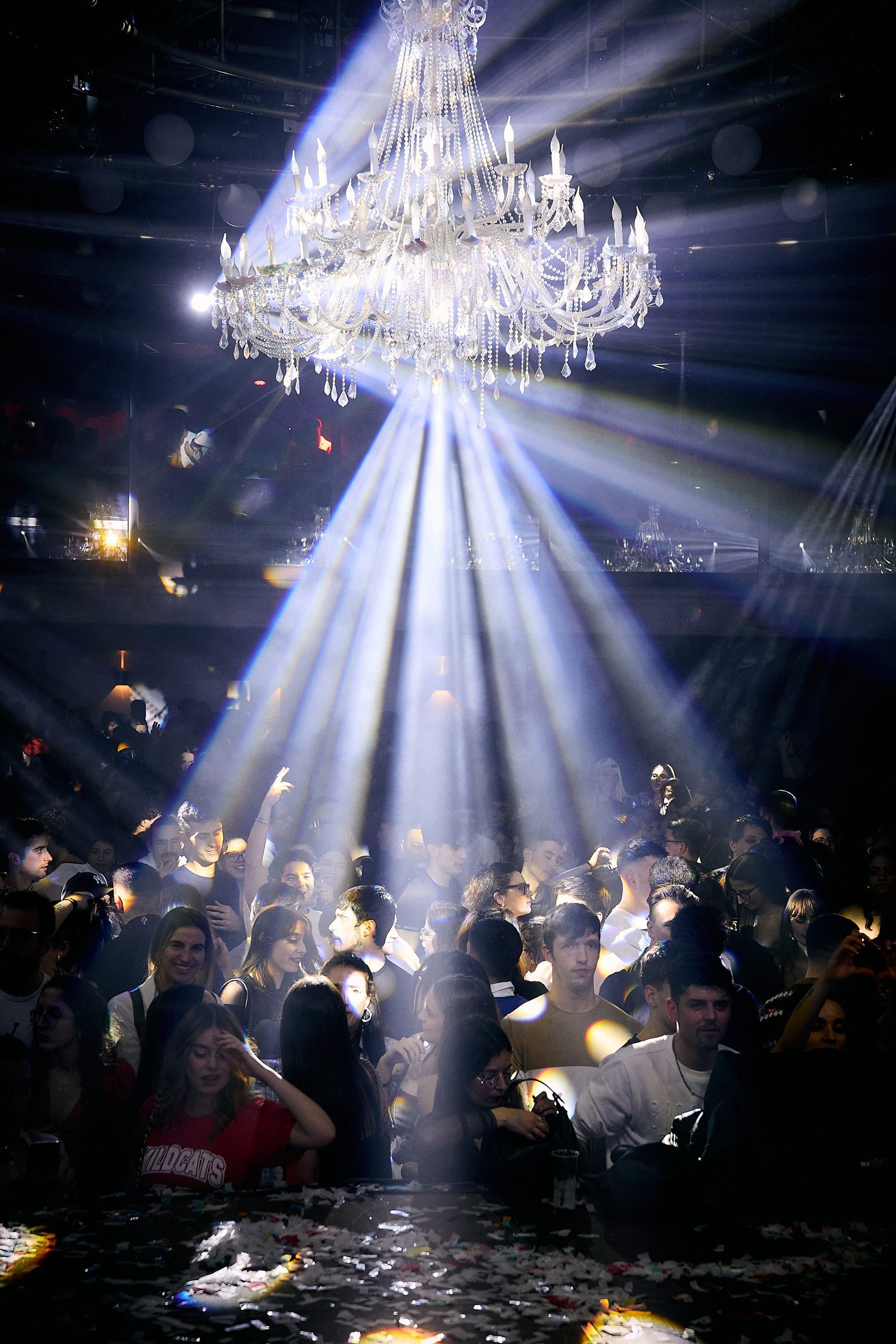 Elegant crystal chandelier illuminates a crowded nightclub dance floor with dramatic light beams streaming down.