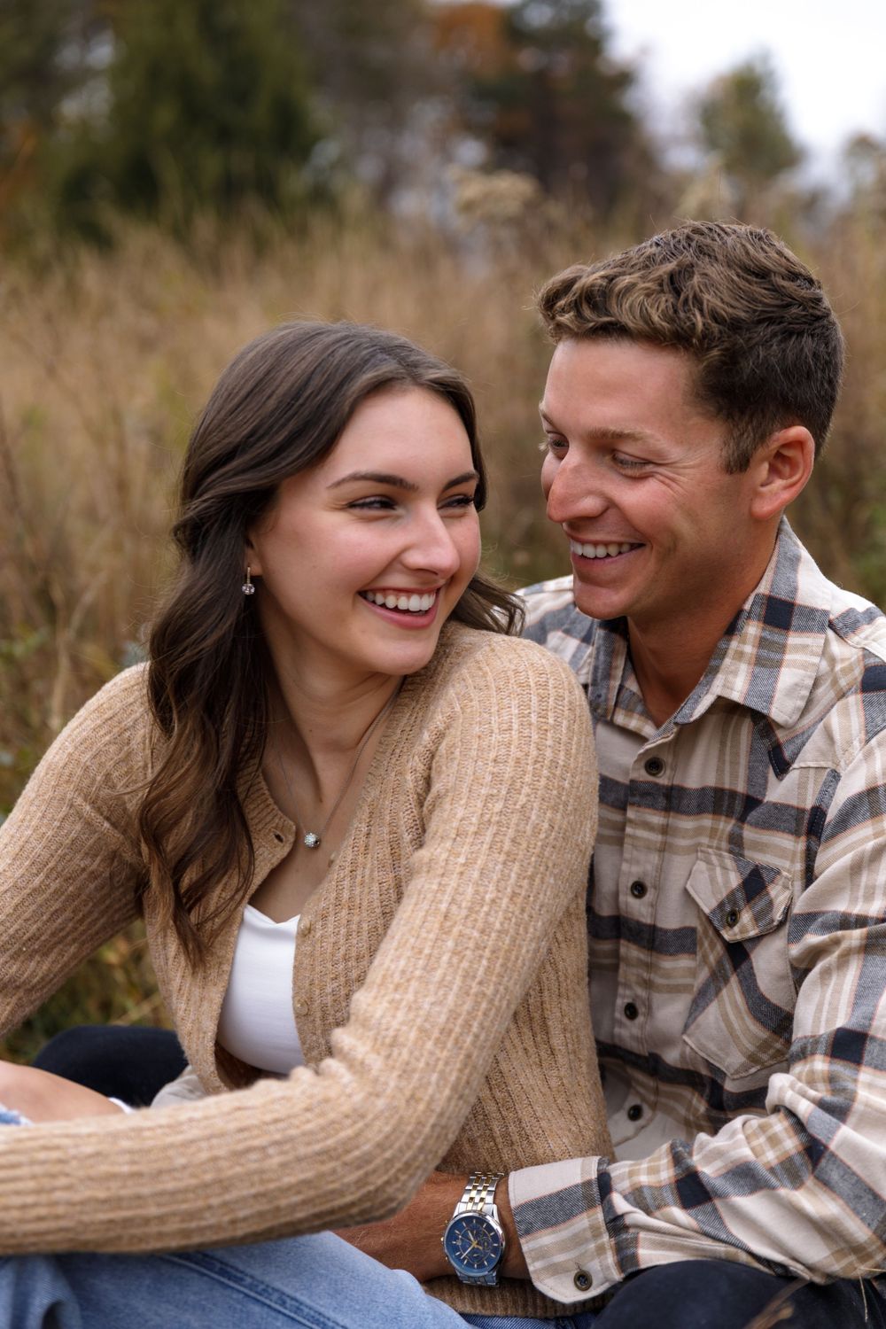 A couple shares laughter while sitting together in a fall field wearing cozy sweater and plaid.