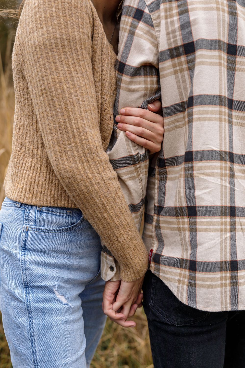 Close up of couple holding hands wearing beige sweater and plaid flannel shirt.