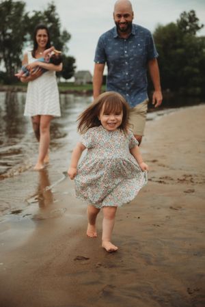 Young child in floral dress running joyfully on wet beach sand while family watches.