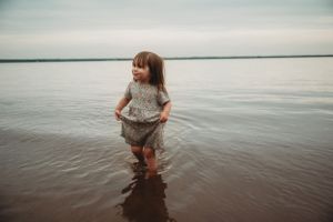 Small child in gray dress playing in shallow water with calm lake horizon in background.