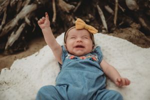 Baby wearing blue outfit and yellow bow lying on white blanket with raised arm.