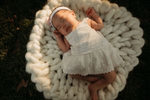 Sleeping baby in white dress on chunky knit blanket outdoors.