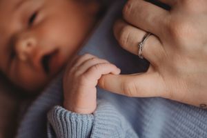 Baby's tiny hand grasping an adult finger wearing a ring against blue knit clothing.