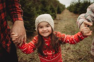 Child in red holiday sweater smiling while holding hands with parents at Christmas tree farm.